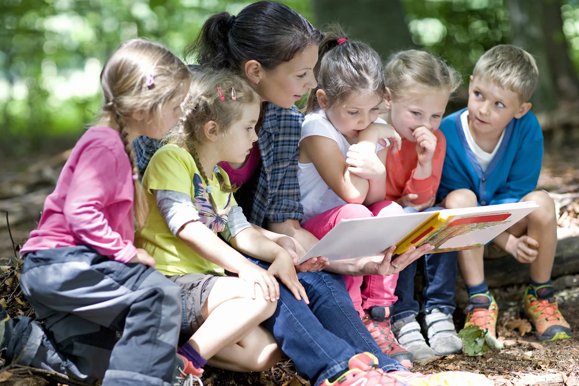 A nursery practitioner reading a picture book with a group of children outdoors